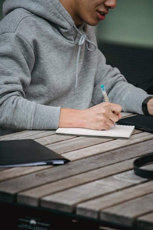 Crop unrecognizable male student in gray hoodie writing notes in notepad while sitting at table and doing homework