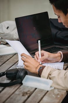 Photo by Armin Rimoldi Young man studying outdoors with a laptop and notebook, focused on his work.