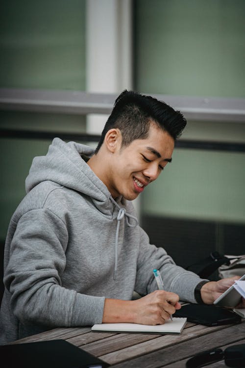 Free Positive young Asian male student taking notes in notepad with tablet in hand while working on project at wooden table Stock Photo