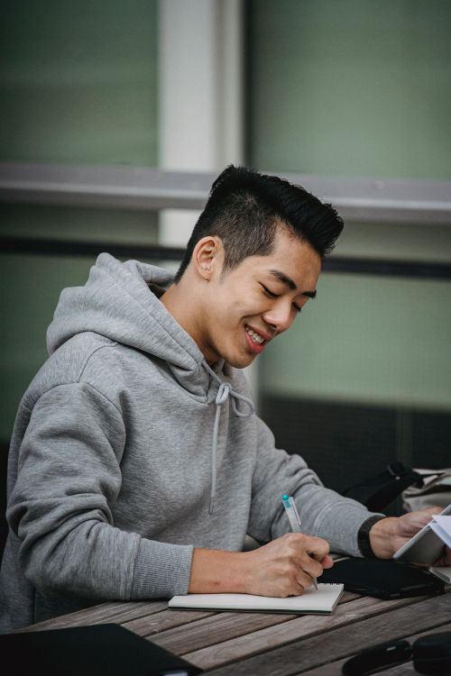 Free Positive young Asian male student taking notes in notepad with tablet in hand while working on project at wooden table Stock Photo