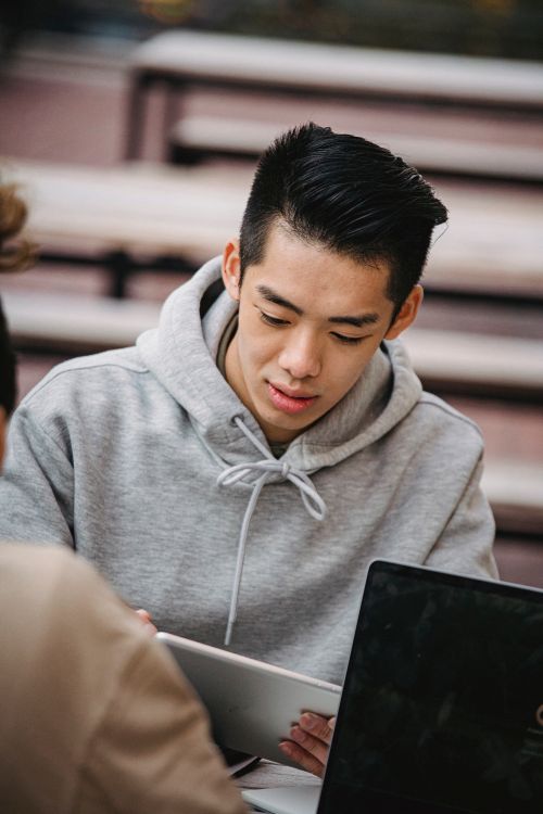 Free Focused young Asian male student in gray hoodie using tablet while working on project together with friend Stock Photo