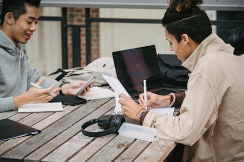 Side view of young multiracial male students in casual clothes sitting at wooden table with gadgets and taking notes while working on project