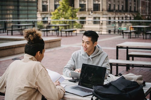 Cheerful young diverse male students in casual wear sitting at wooden table and doing home task together