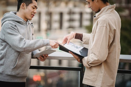 Free Side view of young diverse male students in casual clothes standing on street with tablet and papers in folder and speaking about home task Stock Photo