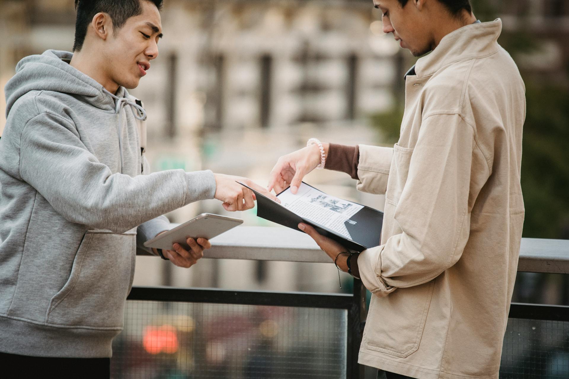 Side view of young diverse male students in casual clothes standing on street with tablet and papers in folder and speaking about home task