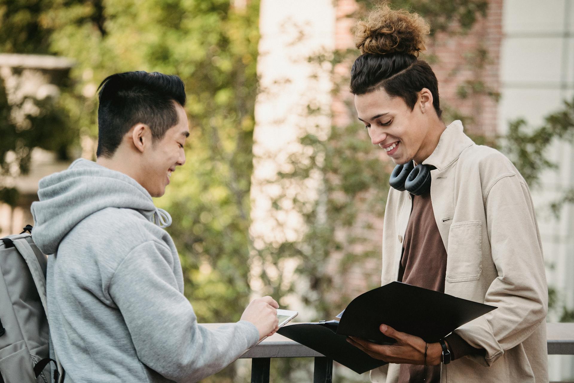 Side view of positive young multiracial male students in casual outfits with folder and tablet talking about research