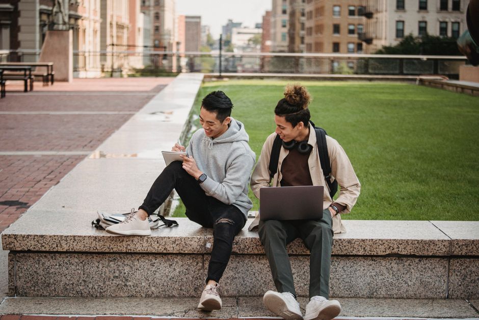 Full body of positive young multiracial male students in casual clothes sitting on stone border with netbook and notebook while speaking about research Full body of positive young multiracial male students in casual clothes sitting on stone border with netbook and notebook while speaking about research