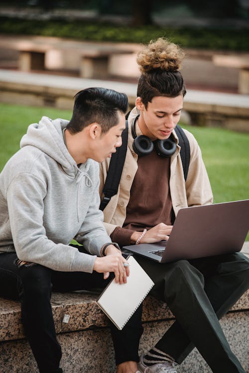 Young multiracial male friends in casual clothes sitting on stone border and browsing internet on laptop while doing research