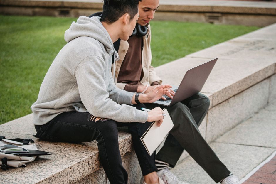 Side view of focused young male students in casual outfits sitting on stone border and surfing internet on laptop while discussing and working on project Side view of focused young male students in casual outfits sitting on stone border and surfing internet on laptop while discussing and working on project