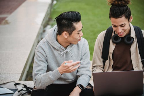 Cheerful young multiracial male friends in casual outfits sitting on stone border and doing and discussing research using laptop