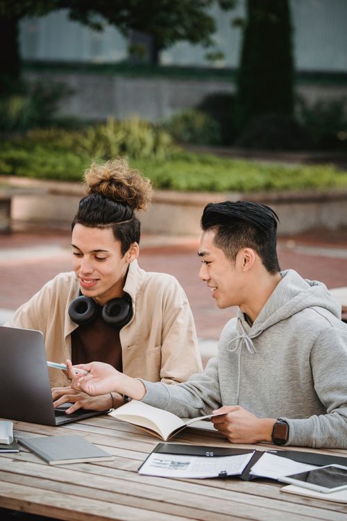 Free Positive young multiracial male students in casual wear sitting at table and working on research with laptop and notebook Stock Photo