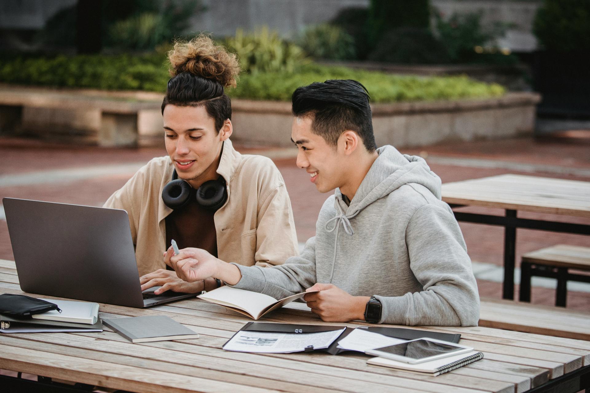 Two young men studying and collaborating outdoors on a campus project, using laptops and notes.