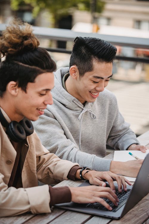 Happy young multiracial male students in casual clothes sitting at table while working on homework together