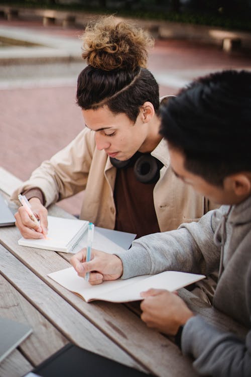 Concentrated male students in casual clothes taking notes in notebooks while preparing for exams together in university campus park