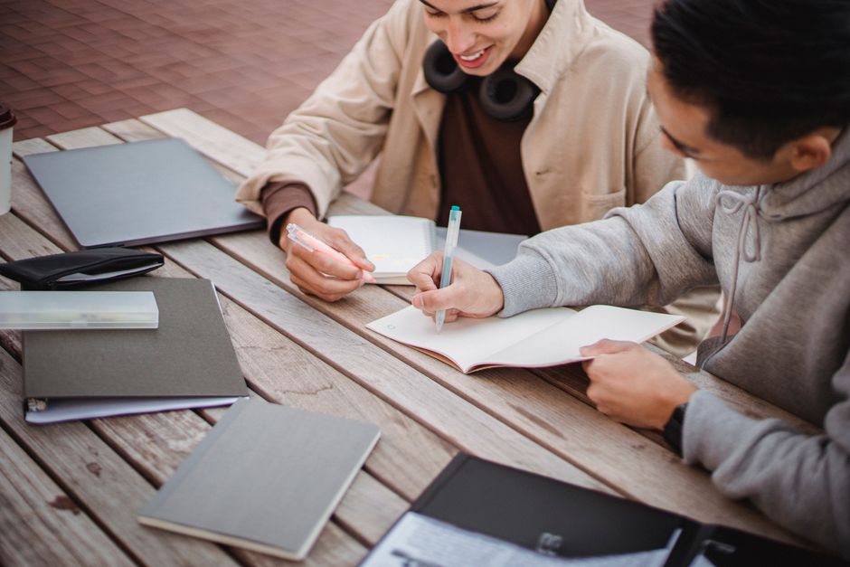 High angle crop happy male in casual clothes taking notes in copybook and doing homework together on campus terrace High angle crop happy male in casual clothes taking notes in copybook and doing homework together on campus terrace
