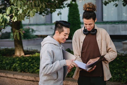 Smiling multiracial male students wearing casual clothes reading article in textbook before seminar while standing in campus park