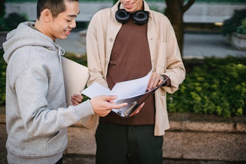 Crop smiling diverse male students wearing casual clothes looking through pages in folder before seminar while standing in lush campus park