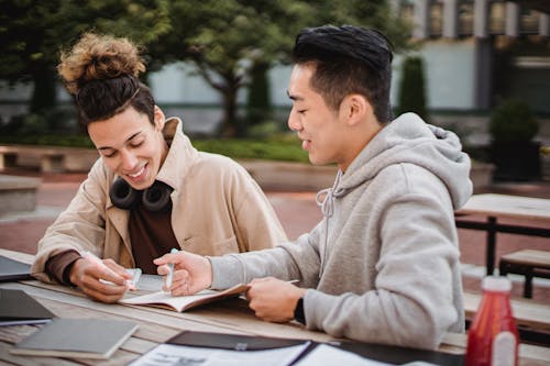 Free Cheerful multiracial male students wearing casual outfit working on college project together while sitting at wooden table in campus park Stock Photo