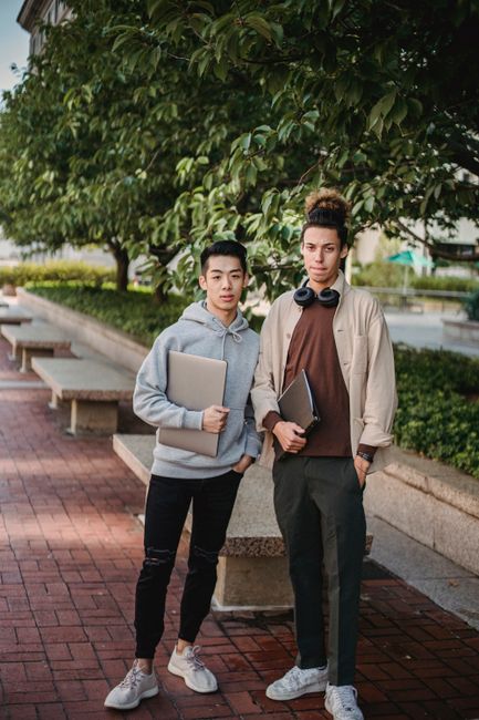 Content multiethnic male students in casual clothes with netbook and folder standing together in verdant sunny park Content multiethnic male students in casual clothes with netbook and folder standing together in verdant sunny park