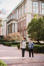 Full body content young male friends with backpacks strolling and chatting on paved sidewalk near modern brick building on sunny weather