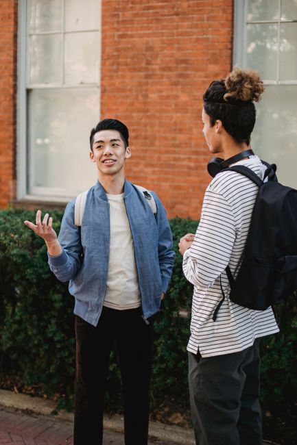 Content young multiracial male friends wearing casual outfits with backpacks having conversation and standing on paved sidewalk on clear day Content young multiracial male friends wearing casual outfits with backpacks having conversation and standing on paved sidewalk on clear day