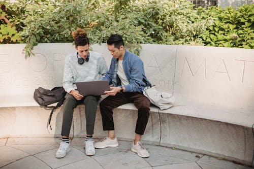 Full length positive multiracial male friends in casual outfits with backpacks browsing modern netbook while sitting on stone bench in green park