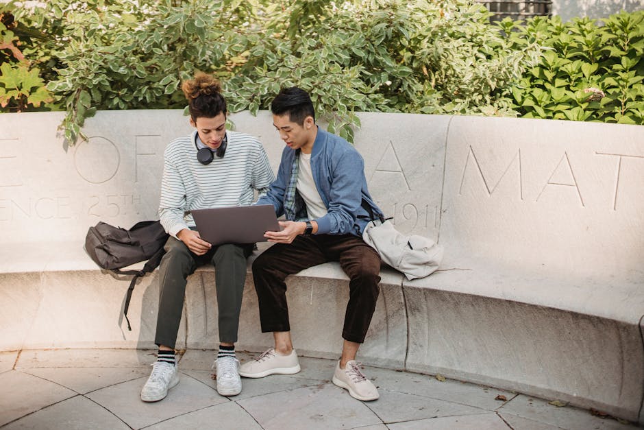 Full length positive multiracial male friends in casual outfits with backpacks browsing modern netbook while sitting on stone bench in green park