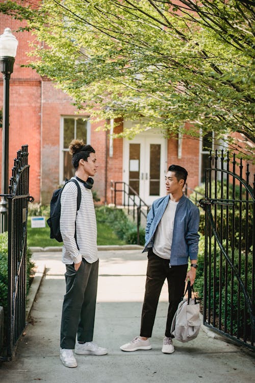 Full length positive young multiracial friends in casual clothes with backpacks talking and looking at each other while standing on street outside brick building