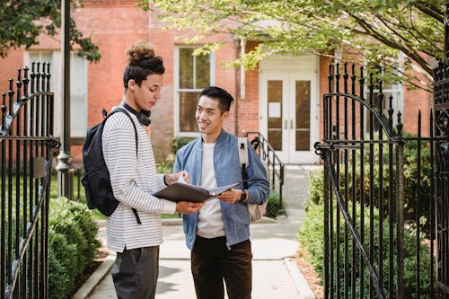 Cheerful diverse male classmates with paper document in folder standing close and discussing task at university entrance