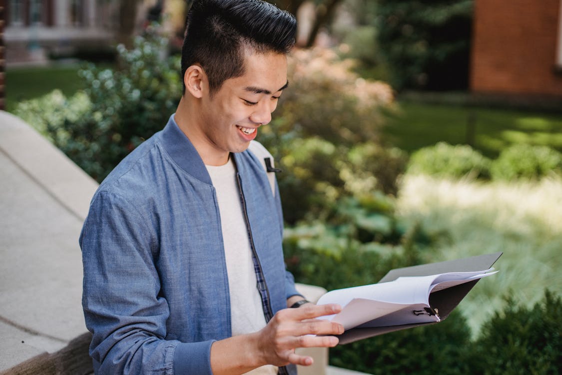Side view of positive Asian male learner looking at papers with project with smile