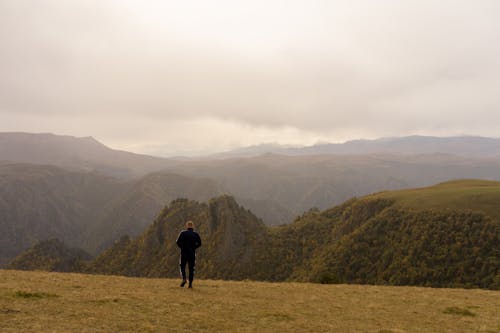 Back view of unrecognizable man walking on grassy ground surrounded by peaks of mountains in cloudy day