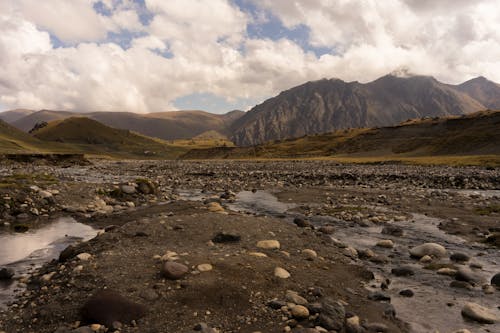 Free Dried river with rocky coast located near green hills against high mountain in cloudy day Stock Photo