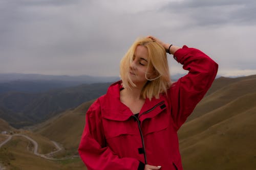 Cheerful young female with short blond hair touching head and standing with closed eyes on top of hill in cloudy day