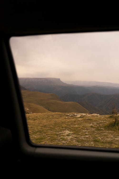 Picturesque view of mountain ridge and dry grassy hills under cloudy sky through car window