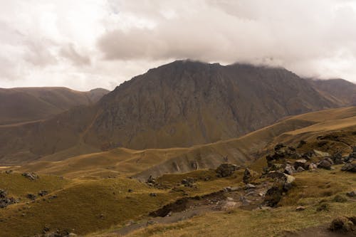 Breathtaking landscape of highland terrain with grass and stones near rocky mountain under cloudy sky