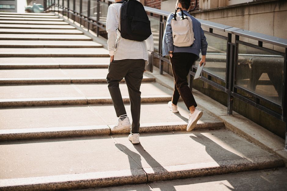 Two friends walking up city steps carrying backpacks, enjoying a sunny day.