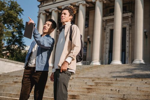 Free From below of Asian male student with folder pointing away while standing near friend attentively looking away Stock Photo