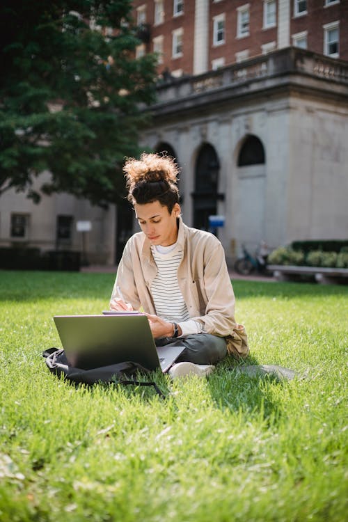 Concentrated male student sitting on green grass while browsing laptop and writing information in notepad