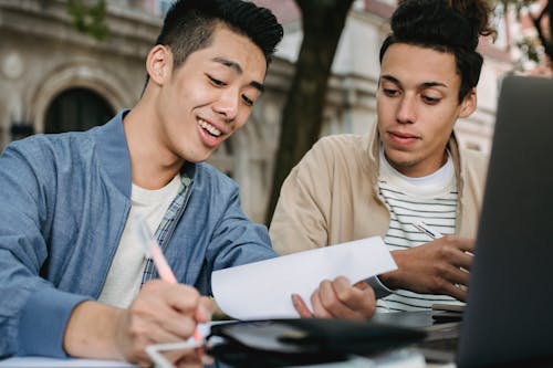 Smiling ethnic male learner writing in document while doing homework assignment with classmate in park