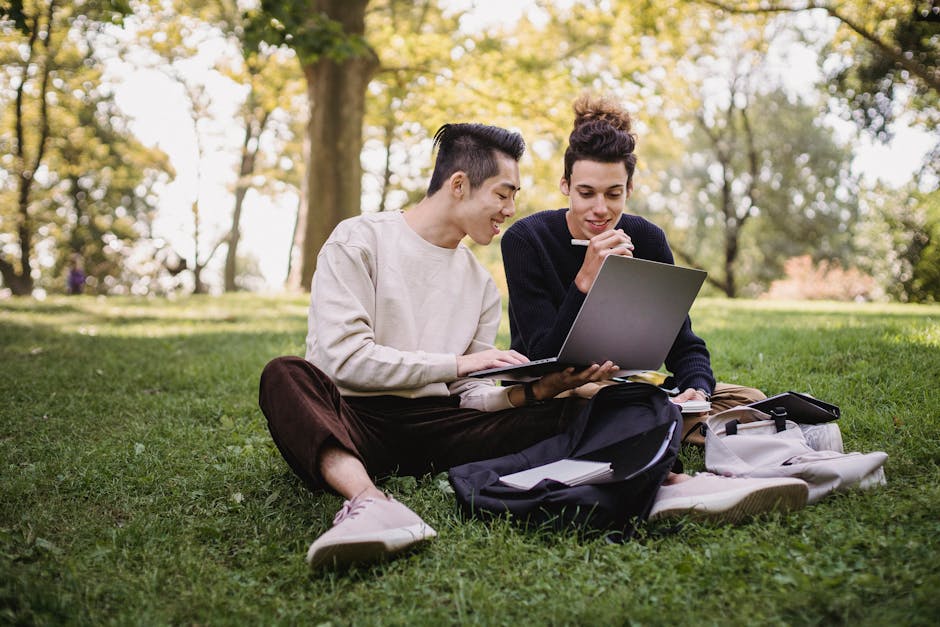 Two young men studying on a laptop while sitting on grass in a park. Two young men studying on a laptop while sitting on grass in a park.