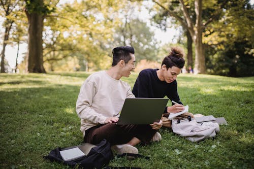 Cheerful Asian male browsing laptop while studying with friend taking notes in notebook in lawn