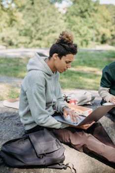 A young adult studying on a laptop outdoors in a park setting.