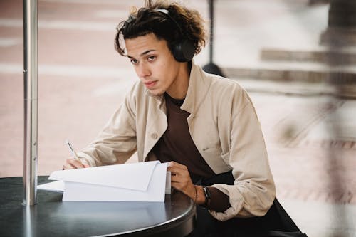 Concentrated male student in wireless headphones sitting at table and writing answers in document during outdoor studies