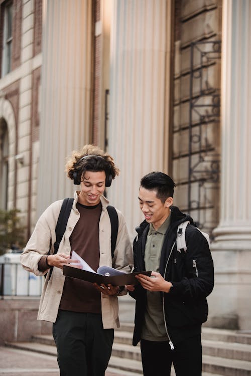 Smiling multiracial classmates in casual wear discussing report together standing near  historic university building