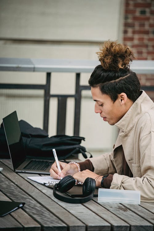 Side view of young ethnic male with stylish haircut taking notes at table with headphones and laptop