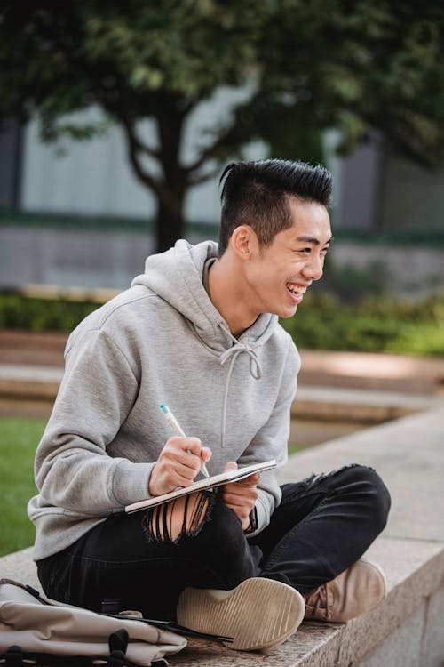 Full body side view of happy young ethnic man in trendy outfit sitting on stone fence with crossed legs and writing information in notebook with pen in park
