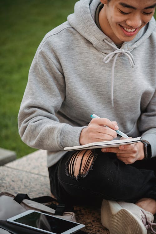 Ethnic man sitting on fence and writing information in notebook