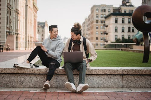 Full body of young multiracial male friends in casual clothes sitting on stone border in park and working on netbook while making notes in notepad in daytime