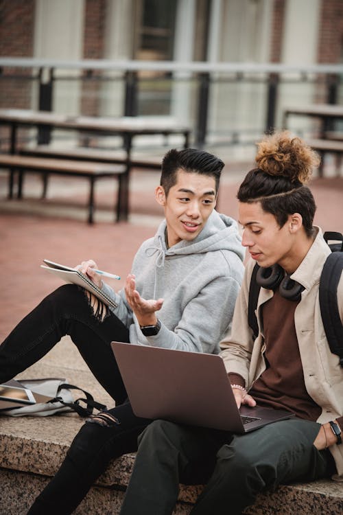 Free Young multiethnic male students in casual outfit sitting on stone border with computer and taking notes in notebook near tablet in daylight Stock Photo