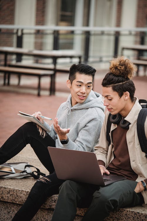 Young multiethnic male students in casual outfit sitting on stone border with computer and taking notes in notebook near tablet in daylight